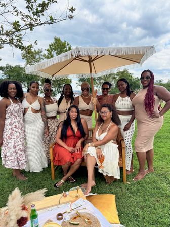 Lakeside park picnic with a group of women in summer dresses smiling under a fringed umbrella, relaxing around a low picnic spread of plates, drinks and cushions on green grass.
