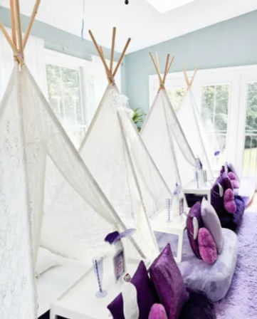 Sunlit indoor kids' sleepover setup with white lace teepees, small white side tables and plush purple and lavender cushions arranged on a soft rug by large windows
