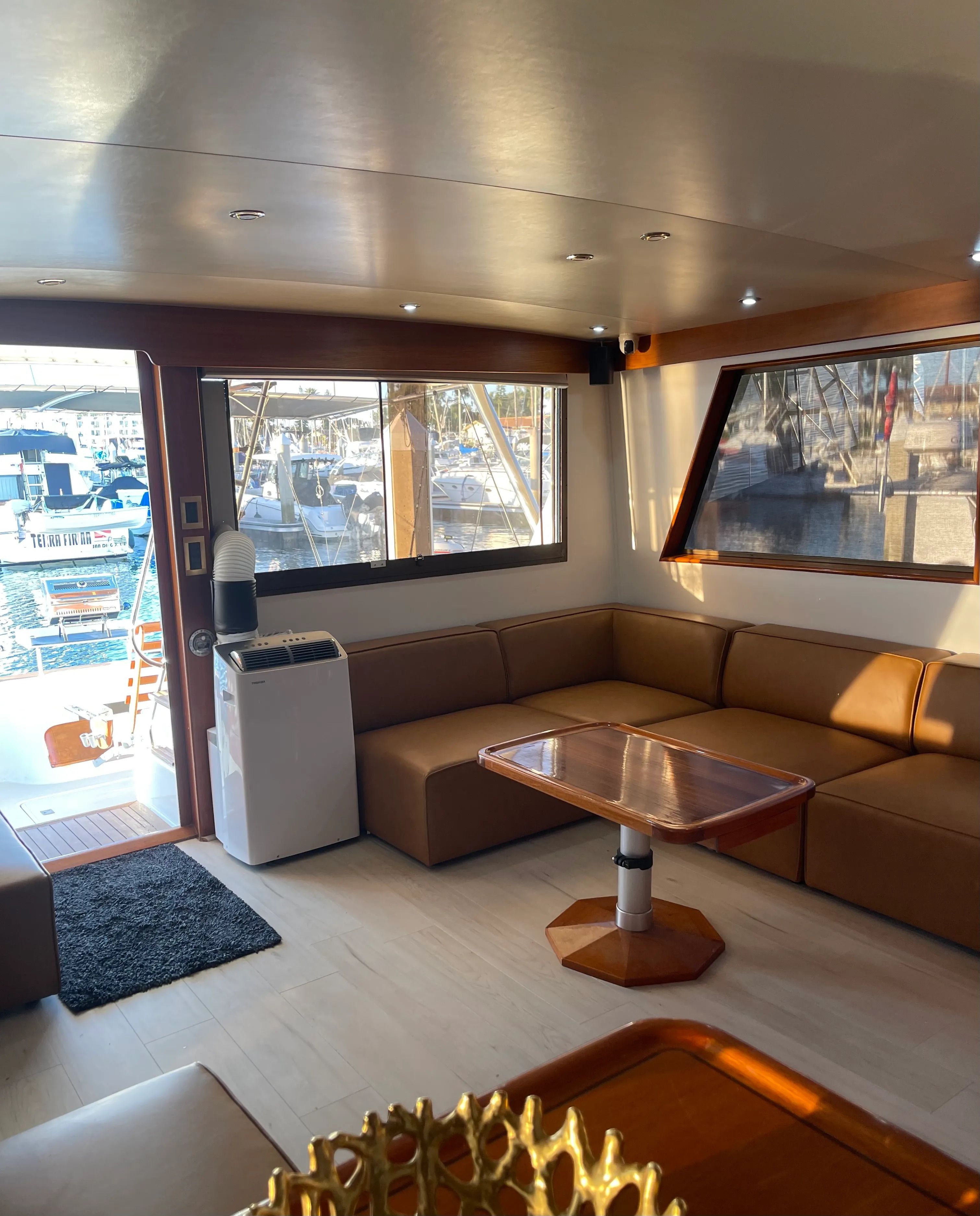 Sunlit yacht salon interior with tan leather L-shaped seating, wooden table, recessed ceiling lights and large windows offering a marina view with docked boats and masts.
