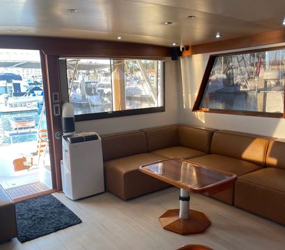 Sunlit yacht salon interior with tan leather L-shaped seating, wooden table, recessed ceiling lights and large windows offering a marina view with docked boats and masts.