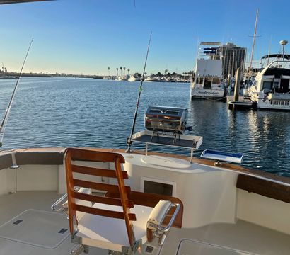 Sunlit boat deck with wooden captain's chair, fishing rods and a grill overlooking a calm marina of yachts, palm trees and clear blue sky.