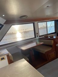 Sunlit yacht dinette with wood-trimmed table and cushioned benches, large windows framing a marina and palm tree silhouettes at sunset.