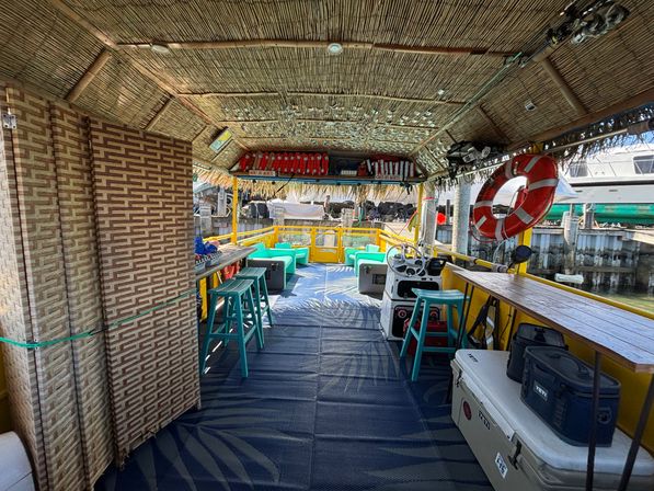 Thatched-roof tropical boat docked at a marina with yellow railings, turquoise bar stools and cushioned seating, a helm with steering wheel, red life ring and stacked life jackets, coolers and wooden bar counter.
