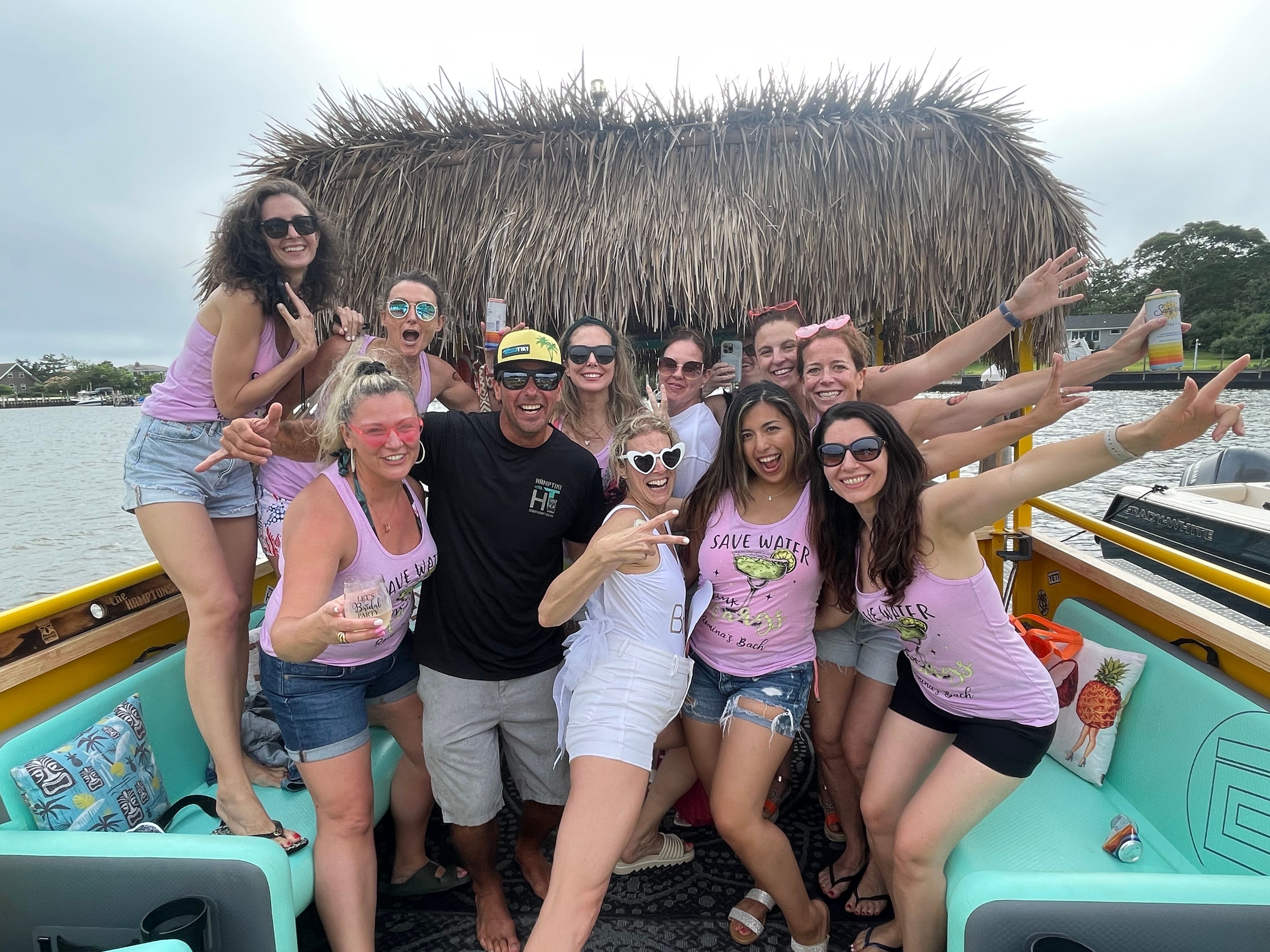 Group of friends partying on a tiki-roof boat in a coastal inlet, wearing matching pink “Save Water” tanks, sunglasses and holding drinks