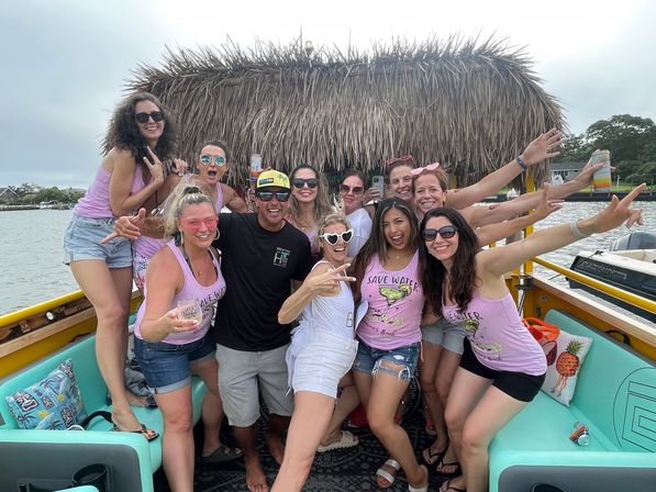 Group of friends partying on a tiki-roof boat in a coastal inlet, wearing matching pink “Save Water” tanks, sunglasses and holding drinks