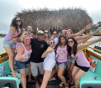 Group of friends partying on a tiki-roof boat in a coastal inlet, wearing matching pink “Save Water” tanks, sunglasses and holding drinks