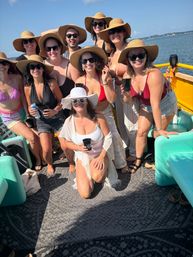 Group of friends in swimsuits and wide-brim sun hats smiling and holding drinks on a sunny pontoon boat with coastal waters and shoreline in the background