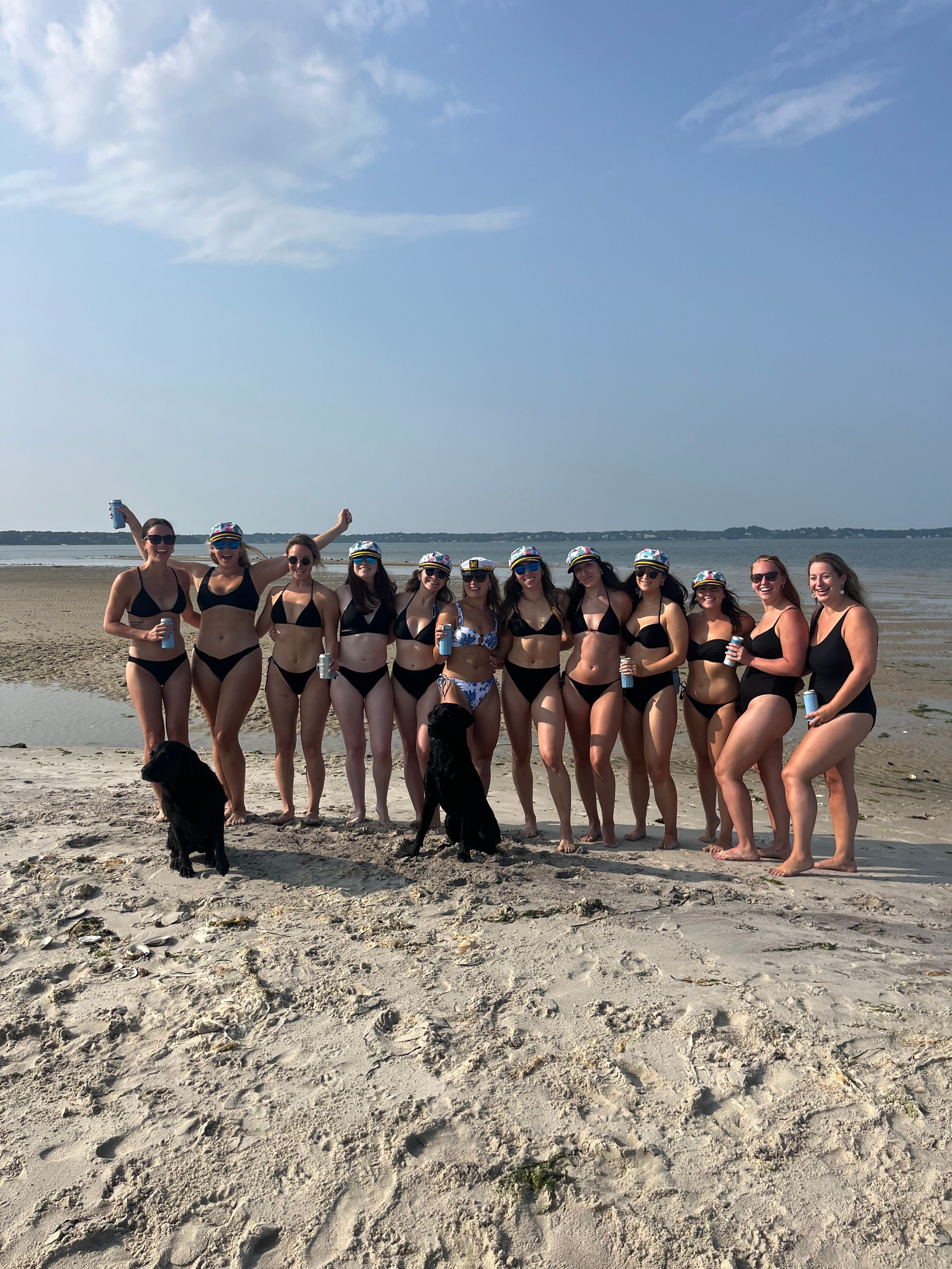 Group of women in swimsuits and colorful captain-style hats lined up on a sandy coastal beach at low tide, holding drinks with two black dogs in front and calm sea and blue sky behind.