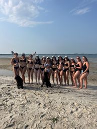 Group of women in mostly black swimsuits and colorful captain hats posing with two black dogs on a sunny sandy beach by the ocean at low tide