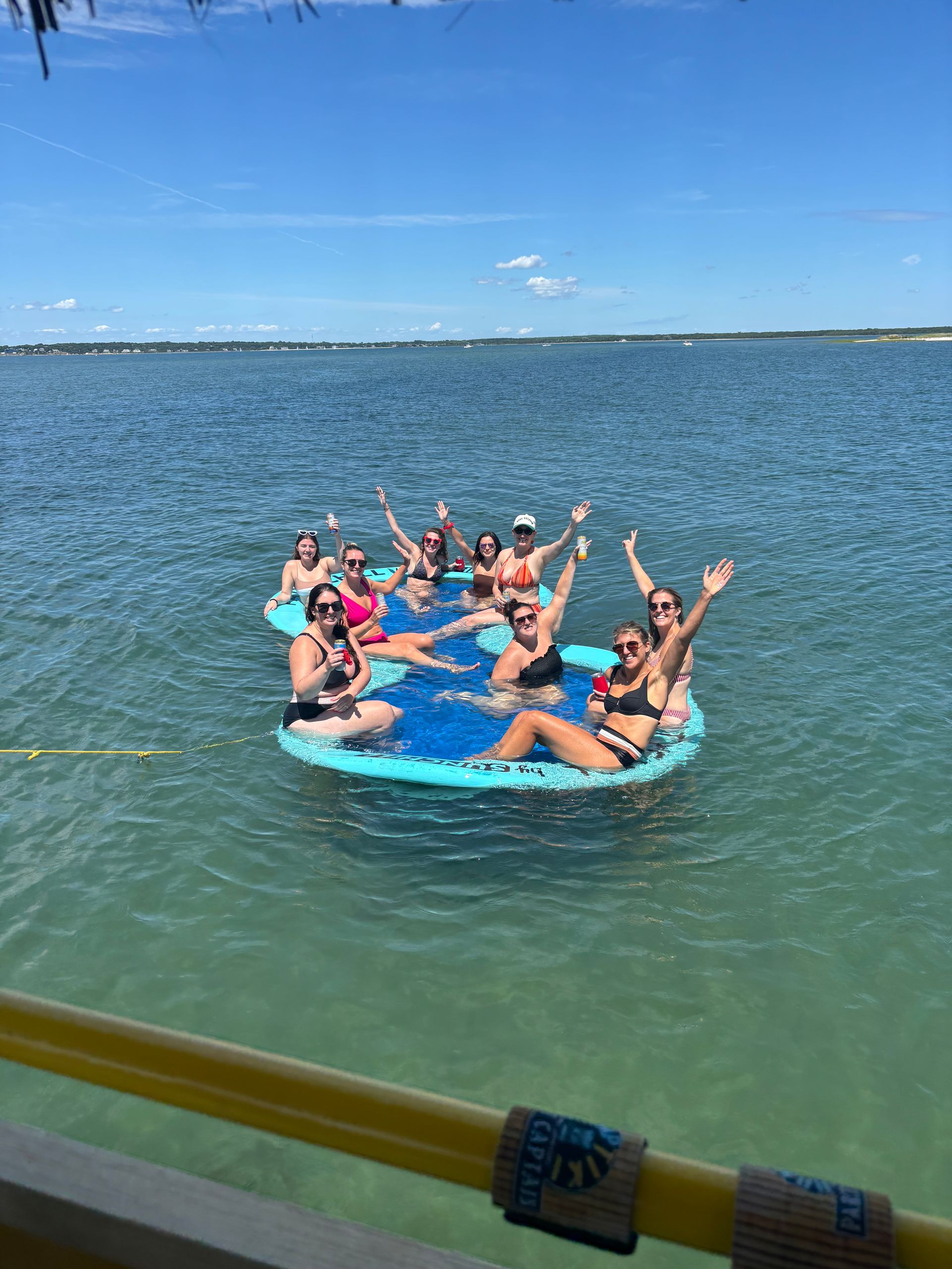 Group of women relaxing on a large blue floating raft in a calm coastal bay, waving and holding drinks under a clear summer sky, seen from a nearby boat.