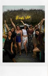 Friends celebrating a bachelorette party on a tropical tiki-roof boat, bride in a captain's hat and white outfit posing with women in summer clothes under a gold "bride tribe" sign.