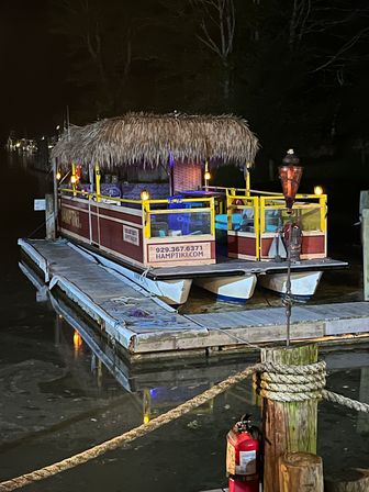 Tiki-style pontoon boat with thatched roof and colorful seating, lit by torches and string lights, docked at a wooden marina pier at night with calm water reflections and a fire extinguisher on a rope-wrapped post.