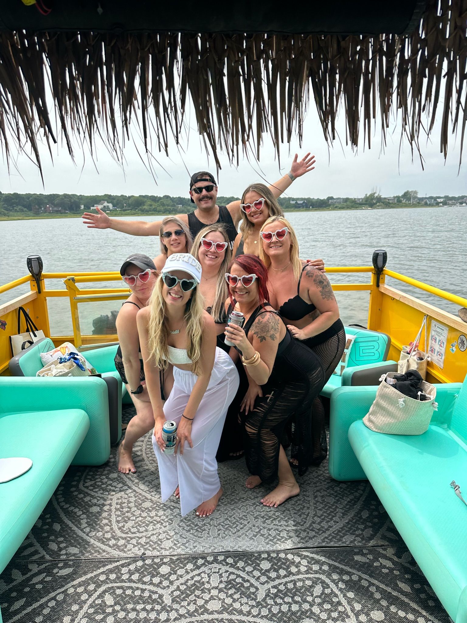 Group of friends on a bright yellow pontoon boat under a thatched tiki roof on a calm lake, wearing heart-shaped sunglasses and summer outfits; turquoise seats and patterned deck mat visible.