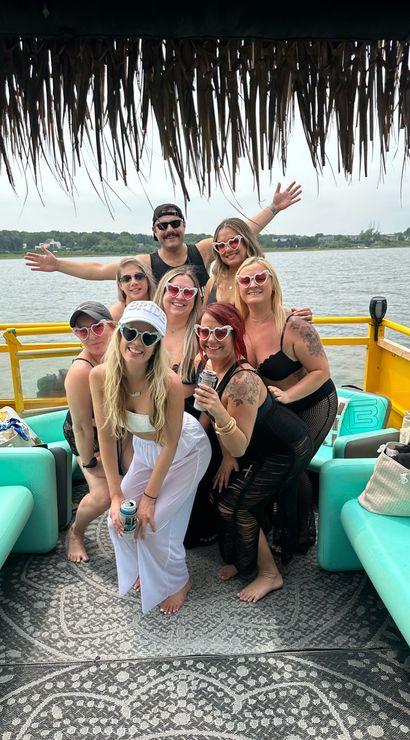 Group of friends on a bright yellow pontoon boat under a thatched tiki roof on a calm lake, wearing heart-shaped sunglasses and summer outfits; turquoise seats and patterned deck mat visible.