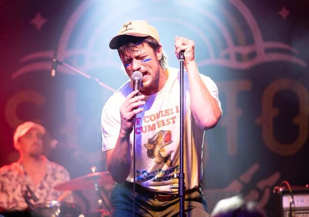 Energetic singer in a baseball cap and blue face paint gripping a microphone on a colorful stage, wearing a vintage fish graphic T-shirt with a drummer blurred behind him.
