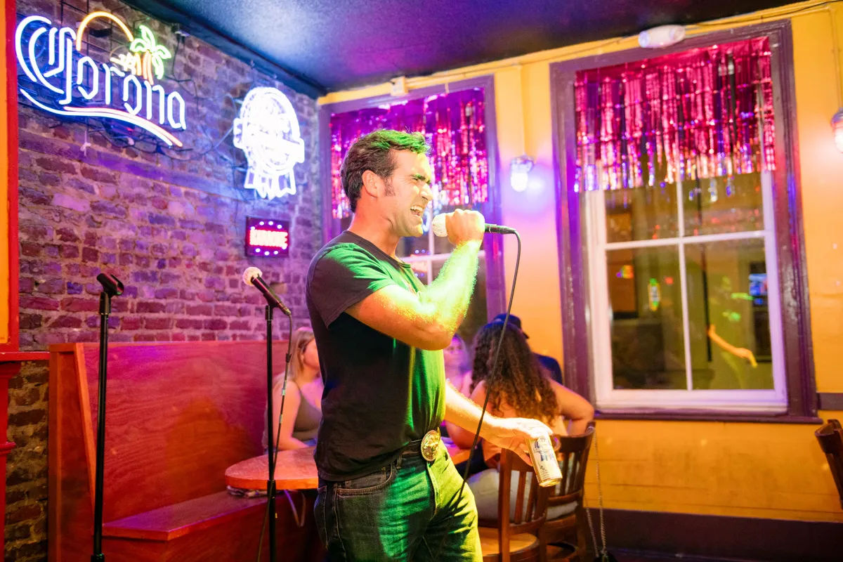 Man passionately singing into a microphone at a colorful karaoke bar, holding a beer can with neon beer signs, exposed brick wall, glittery window curtains and seated patrons in the background — lively nightlife scene.