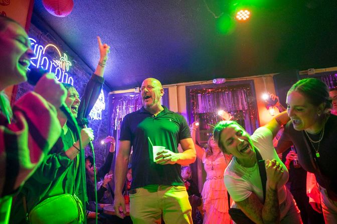 Friends singing and cheering in a crowded karaoke bar under colorful neon and green stage lights, with a man holding a drink at center and lively nighttime atmosphere