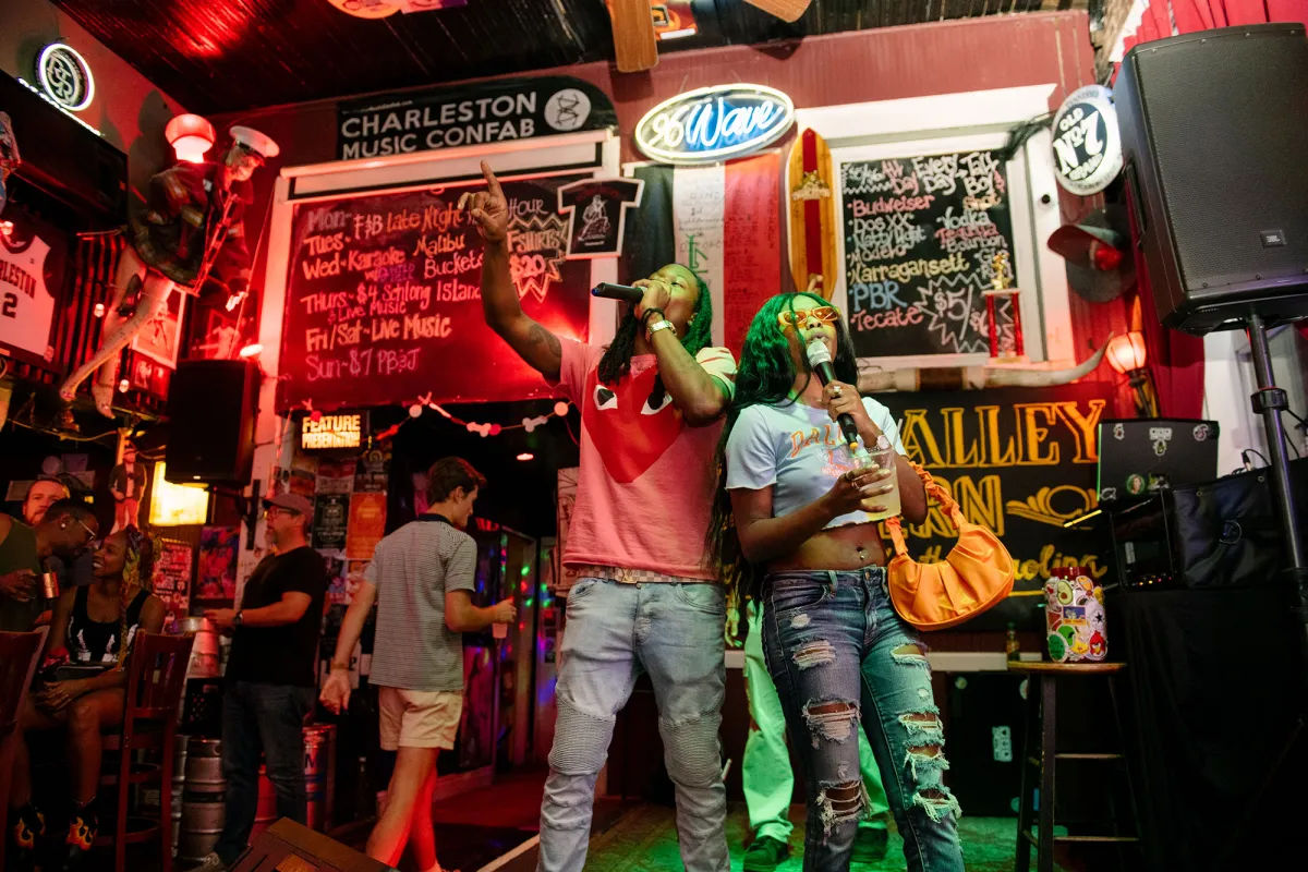 Two friends singing onstage at a neon-lit Charleston bar karaoke night, man in a pink tee pointing up and woman in ripped jeans holding a drink with chalkboard specials and colorful signage behind them.