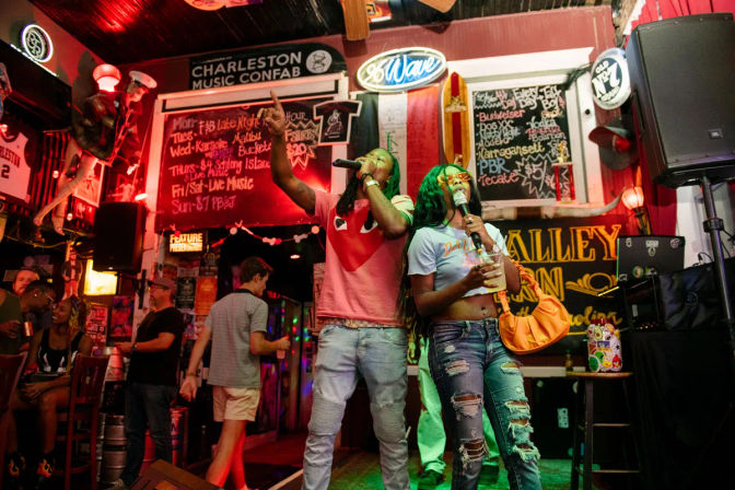Two friends singing onstage at a neon-lit Charleston bar karaoke night, man in a pink tee pointing up and woman in ripped jeans holding a drink with chalkboard specials and colorful signage behind them.