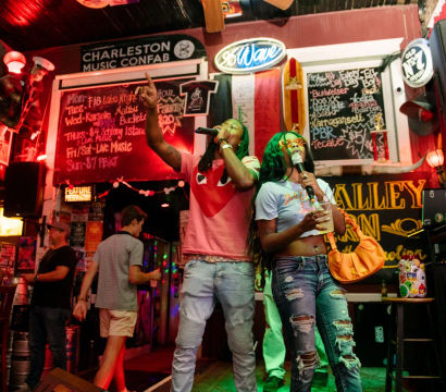 Two friends singing onstage at a neon-lit Charleston bar karaoke night, man in a pink tee pointing up and woman in ripped jeans holding a drink with chalkboard specials and colorful signage behind them.