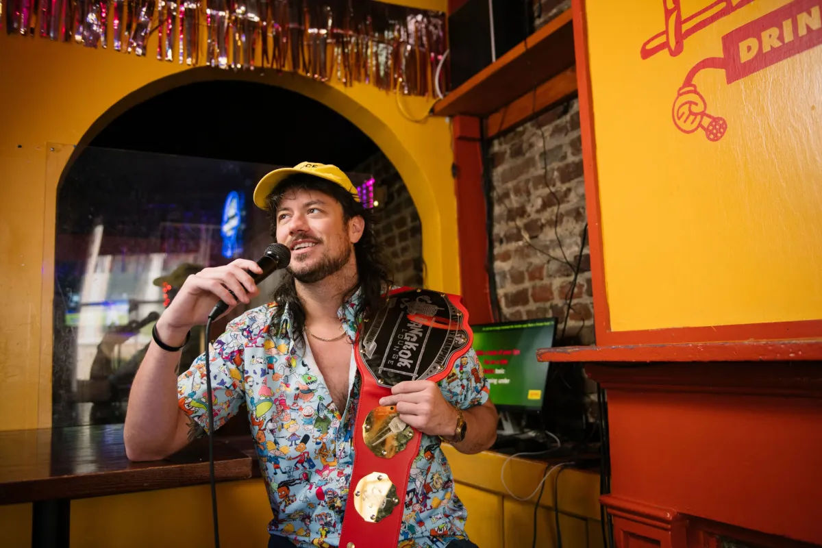 Bearded man in a yellow cap and colorful cartoon shirt smiling while holding a microphone and a red novelty championship belt inside a lively yellow-orange karaoke bar with exposed brick and a karaoke screen.