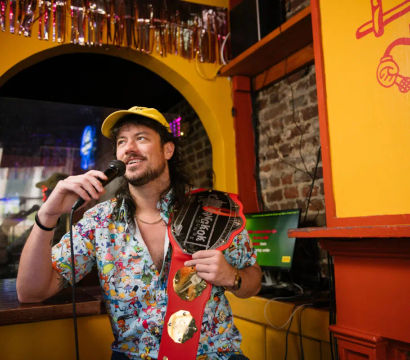Bearded man in a yellow cap and colorful cartoon shirt smiling while holding a microphone and a red novelty championship belt inside a lively yellow-orange karaoke bar with exposed brick and a karaoke screen.