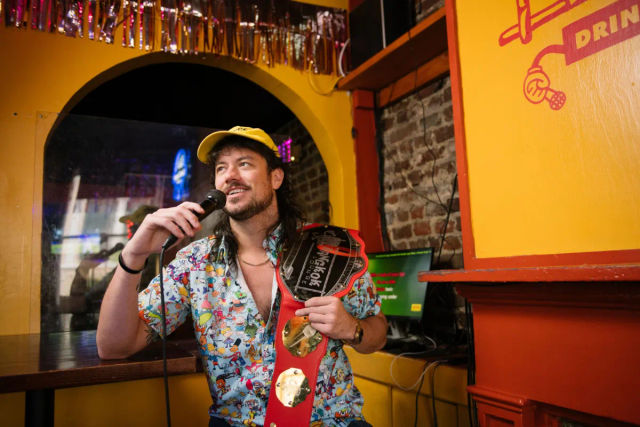 Bearded man in a yellow cap and colorful cartoon shirt smiling while holding a microphone and a red novelty championship belt inside a lively yellow-orange karaoke bar with exposed brick and a karaoke screen.