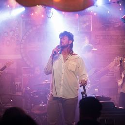 Lead singer with mullet-style haircut holding a microphone on a smoky, purple-lit small venue stage during a live music set, backed by guitarist, bassist and drummer with audience silhouettes in the foreground.