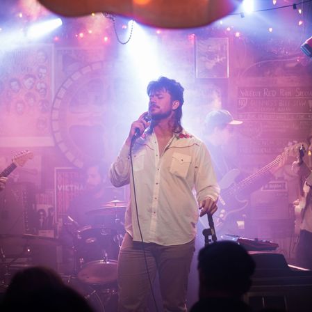 Lead singer with mullet-style haircut holding a microphone on a smoky, purple-lit small venue stage during a live music set, backed by guitarist, bassist and drummer with audience silhouettes in the foreground.