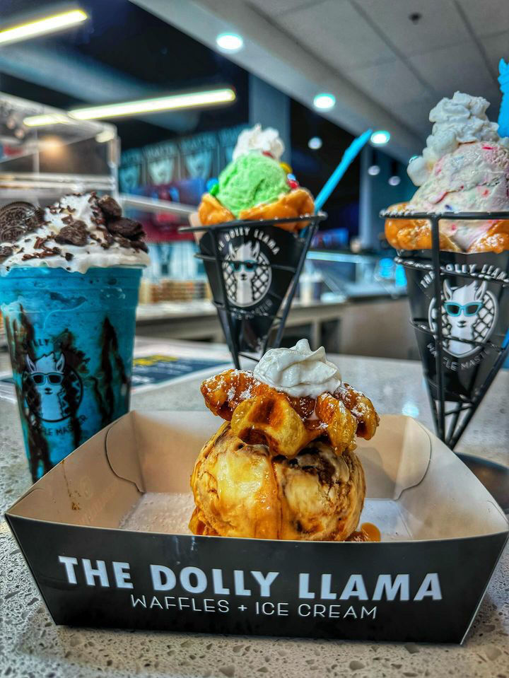 Caramel-drizzled waffle ball topped with whipped cream and ice cream, flanked by colorful scooped cones and a blue cookies-and-cream milkshake on a bright ice cream parlor counter — waffles and ice cream dessert shot.