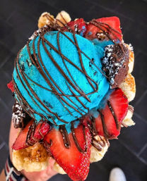 Vibrant blue ice cream scoop on a bubble waffle cone topped with sliced strawberries, banana, brownie bites and chocolate drizzle
