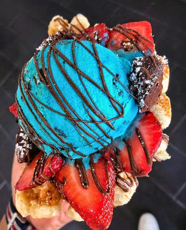Vibrant blue ice cream scoop on a bubble waffle cone topped with sliced strawberries, banana, brownie bites and chocolate drizzle