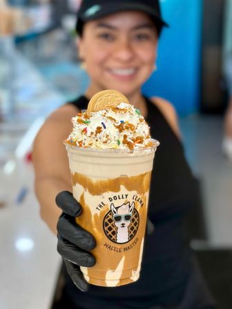 Close-up of a caramel milkshake with whipped cream, colorful sprinkles, cookie and crunchy crumbs, swirled in a clear cup and held by a gloved hand with a blurred smiling server and cafe interior in the background.