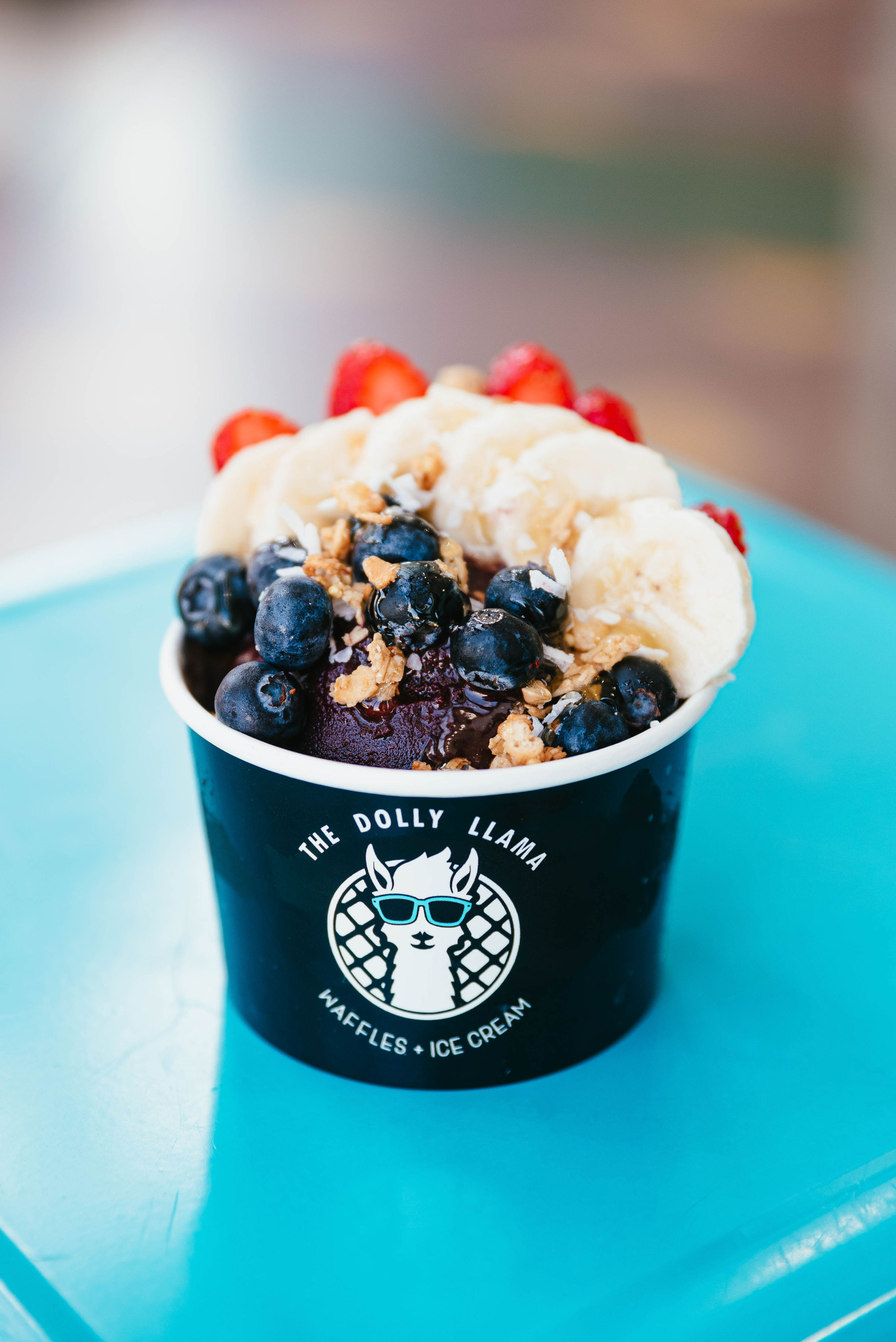 Vibrant acai bowl in a black branded cup on a bright turquoise table, topped with banana slices, fresh blueberries, strawberries and crunchy granola.
