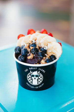 Vibrant acai bowl in a black branded cup on a bright turquoise table, topped with banana slices, fresh blueberries, strawberries and crunchy granola.