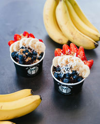 Two acai bowls in black cups topped with banana slices, strawberry fans, blueberries, shredded coconut and granola, set on a dark table with bunches of ripe bananas nearby.