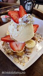 Waffle dessert with a scoop of vanilla ice cream, fresh strawberry slices, banana rounds, toasted almond flakes and powdered sugar on a white plate at a café table.