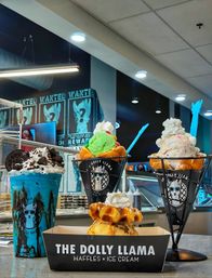 Vibrant dessert display on an indoor ice cream shop counter: two waffle-cone cups with colorful scoops and whipped cream, a blue cookies-and-cream milkshake, and a mini waffle topped with whipped cream.