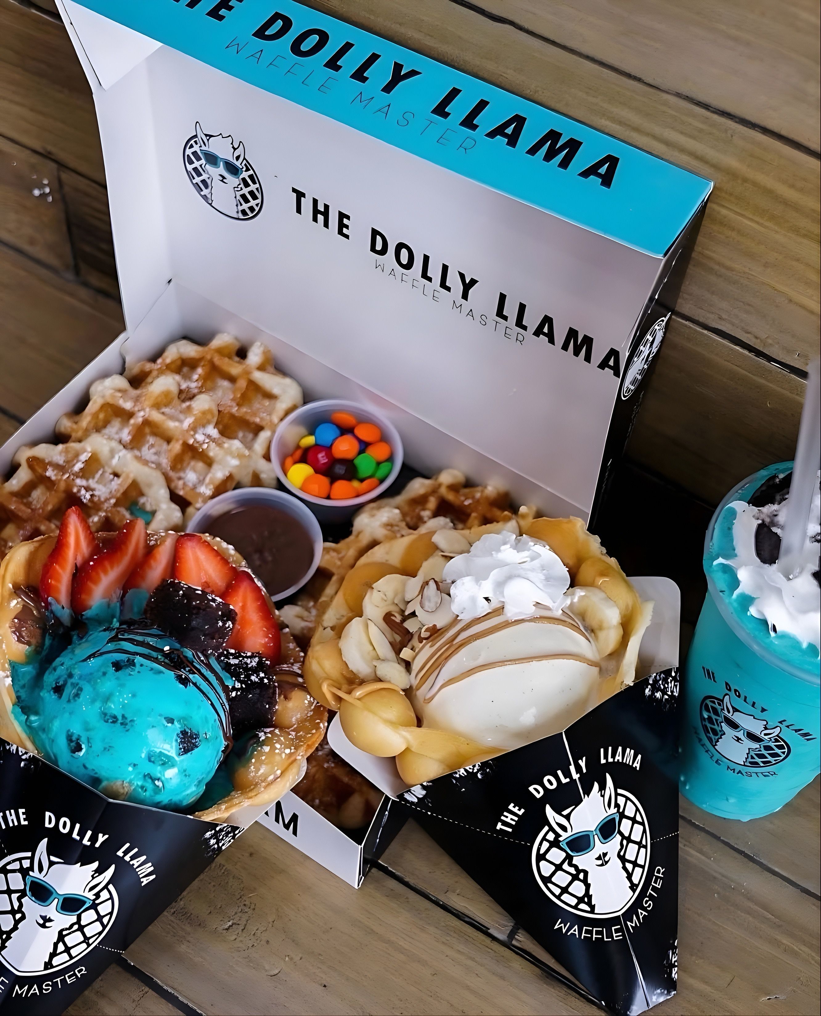 Dessert box with a Belgian waffle and two bubble-waffle cones topped with bright blue ice cream, fresh strawberries, chocolate drizzle, banana slices, vanilla scoop, colorful candy and dipping sauces, plus a turquoise milkshake on a wooden table.