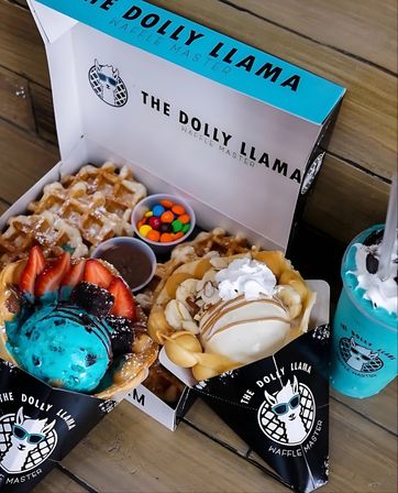 Dessert box with a Belgian waffle and two bubble-waffle cones topped with bright blue ice cream, fresh strawberries, chocolate drizzle, banana slices, vanilla scoop, colorful candy and dipping sauces, plus a turquoise milkshake on a wooden table.