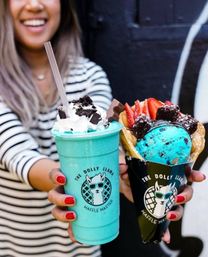 Smiling person in a striped shirt holding a bright turquoise milkshake topped with whipped cream and cookie crumbs beside a bubble waffle cone filled with blue ice cream, sliced strawberries and brownie bites — colorful urban dessert treat.