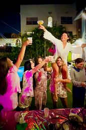 Group of friends at a nighttime backyard party by a pool and palm trees, one person on another’s shoulders raising a cocktail while others cheer and toast under colorful lights.