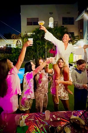 Group of friends at a nighttime backyard party by a pool and palm trees, one person on another’s shoulders raising a cocktail while others cheer and toast under colorful lights.