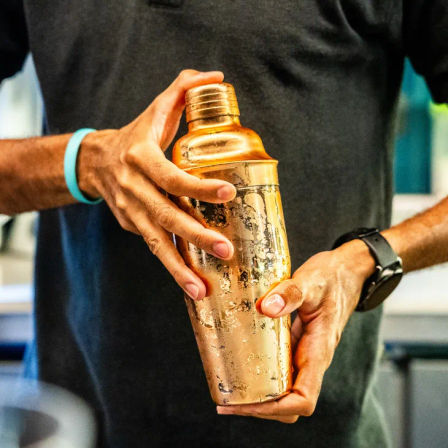 Close-up of hands gripping a worn gold cocktail shaker, ready for mixology in a modern bar setting