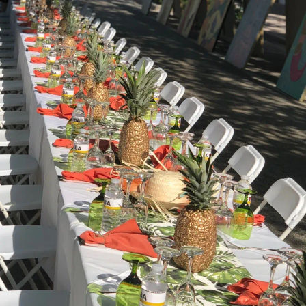 Outdoor tropical-themed long banquet table set for an event with gold pineapple centerpieces, palm-print runner, white folding chairs, orange napkins, green glassware and bottled water
