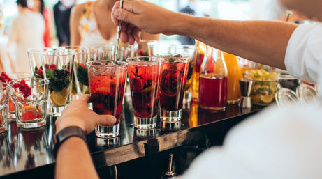 Bartender stirring vibrant red berry cocktails with muddled fruit, mint and lime in tall glasses lined up on a glossy bar counter at a lively event