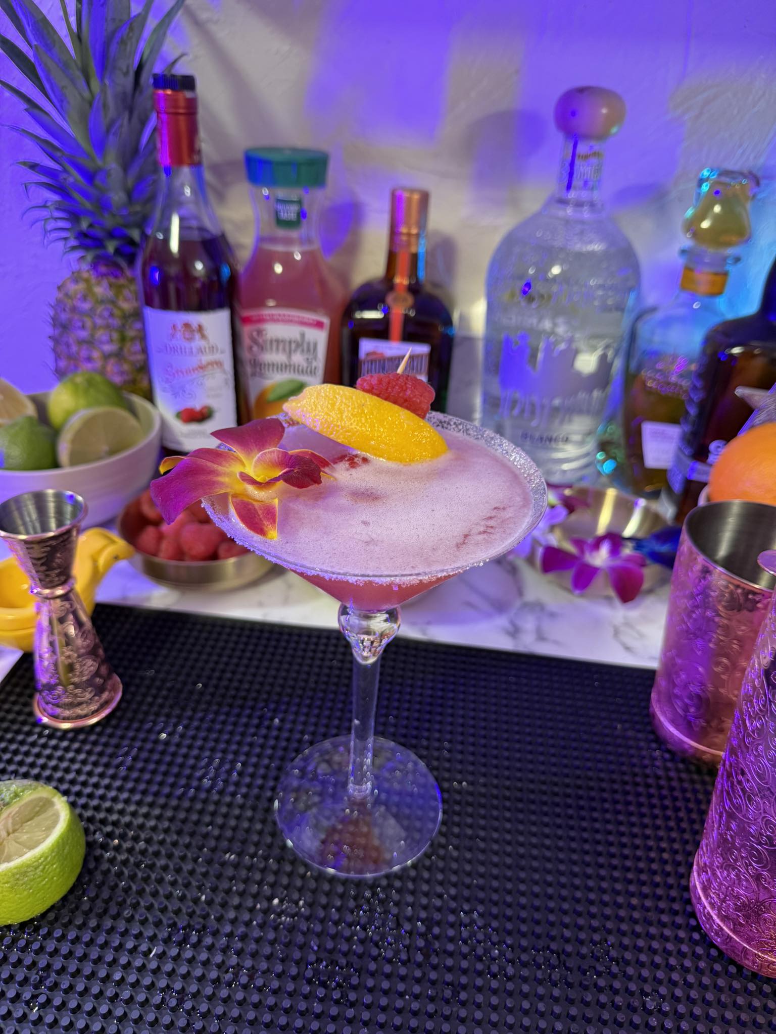 Frosty pink tropical cocktail in a sugar‑rimmed coupe glass, topped with an orange peel, raspberry and purple orchid, staged on a home bar mat with limes, pineapple and liquor bottles under colorful purple-blue lighting.