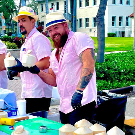 Smiling, tattooed vendor in a pink shirt and straw hat hands a freshly cut coconut at a sunny tropical street market booth