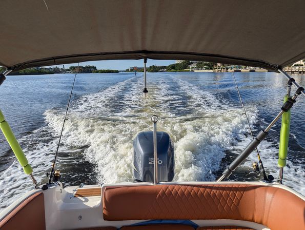 View from the stern of a motorboat under a bimini, showing an outboard engine, two fishing rods and foamy wake on a sunny coastal waterway with shoreline homes.