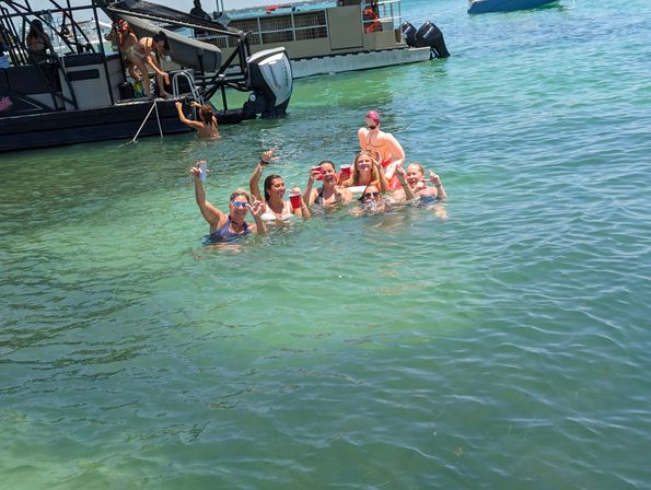Group of people enjoying a sunny boat party in clear turquoise water, smiling and holding red cups near a motorboat ladder.